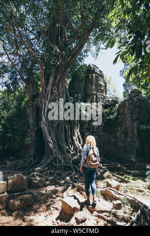 Caucasian donna bionda scoprendo le rovine di Angkor Wat tempio complesso in Siem Reap, Cambogia. Albero che cresce sul tetto del tempio cancello di ingresso arc Foto Stock