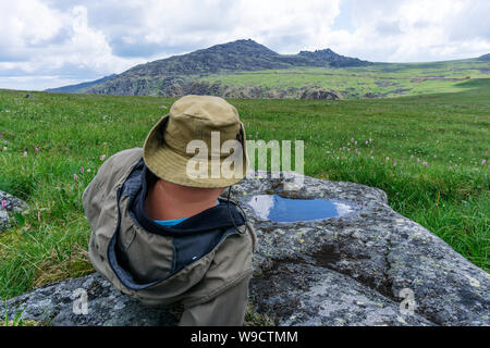 Stanco viaggiatore in montagna - un uomo in un cappello e camp vestiti in appoggio, fissando in distanza, giacente su di un altopiano di montagna Foto Stock
