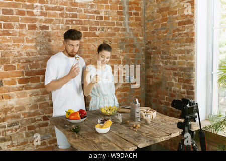 Vlogger e blogger lavoro freelance, food concept. Giovane coppia caucasica cucinare insieme e la registrazione di video live per vlog e social media con telecamera professionale contro un muro di mattoni nella loro cucina. Foto Stock