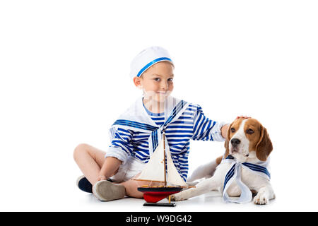 Preschooler sorridente bambino in costume da marinaio con nave giocattolo e cane beagle su bianco Foto Stock