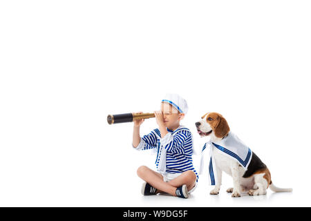 Focalizzato preschooler bambino in costume da marinaio cercando in il cannocchiale mentre è seduto con cane beagle su bianco Foto Stock