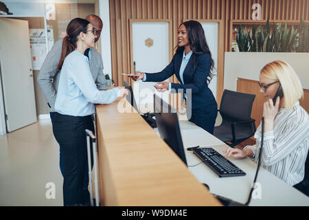 Friendly African American concierge dando due ospiti loro controllare in informazioni mentre si lavora dietro il banco della reception di un hotel Foto Stock