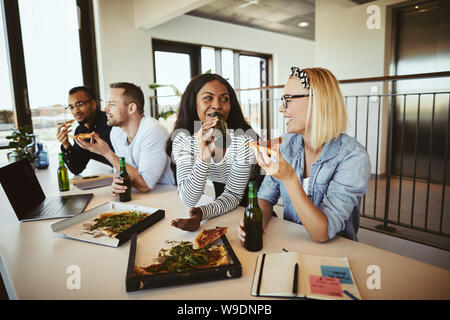 Due diversi giovani imprenditrici sorridente e parlare insieme mentre è seduto con i colleghi in un ufficio tabella dopo i lavori di bere birra e mangiare il piz Foto Stock