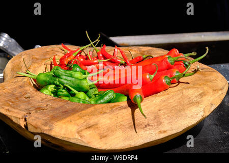 Ciotola di legno sul contatore nero riempito con il verde e il rosso dei peperoncini rossi sul contatore esterno in condizioni di intensa luce di sole Foto Stock