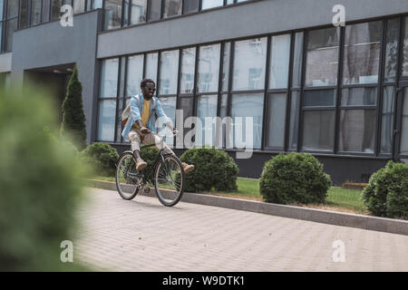 Messa a fuoco selettiva di African American uomo in bicicletta lungo edificio e cespugli verdi Foto Stock