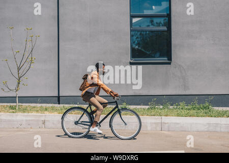 Elegante americano africano imprenditore con zaino Bicicletta Equitazione sulla strada soleggiata Foto Stock