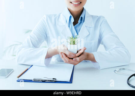 Vista parziale di sorridente medico latina azienda pianta in vaso mentre è seduto al posto di lavoro in clinica Foto Stock