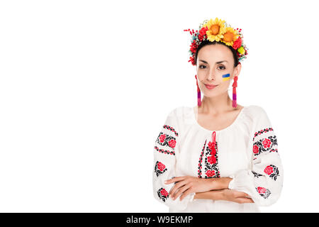 Bruna giovane donna in nazionale ucraino camicia ricamata e ghirlanda floreale con bracci incrociati isolato su bianco Foto Stock