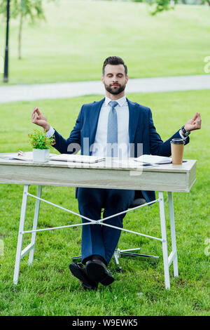 Vista frontale del bel giovane imprenditore nel usura formale la meditazione seduti a tavola con vari office stuff in posizione di parcheggio Foto Stock