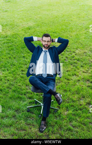 Elevato angolo di visione di imprenditore seduto nella sedia da ufficio in parco con attraversato i GAL e braccia dietro la testa Foto Stock