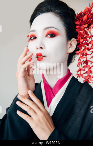 Bella geisha in kimono nero con fiori di colore rosso nei capelli di toccare la faccia isolato su bianco Foto Stock