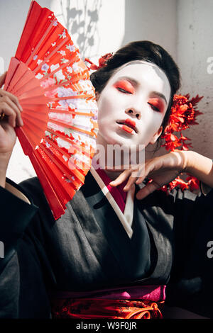 Ritratto di bella geisha con il rosso e bianco specchio tenendo la ventola a mano e toccare la faccia in presenza di luce solare Foto Stock