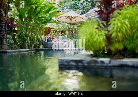 Candid shot della giovane coppia caucasica lounging nella sedia moderna nei pressi di lussuosa piscina di tropical hotel e spa a Bali Foto Stock