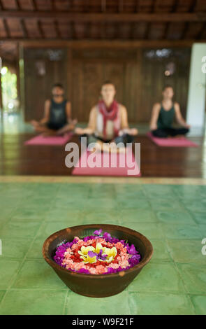 Chiusura del fiore che offre per la preghiera in Indonesiano tradizionale tempio a Bali con tre diverse persone meditando in background Foto Stock