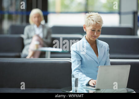 Imprenditrice maturo con corti capelli biondi seduti a tavola e la digitazione sul computer portatile con altra donna seduta in background in ufficio Foto Stock