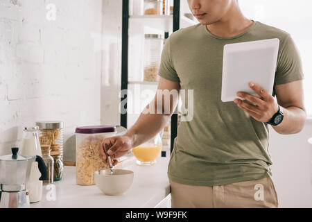 Vista parziale del giovane uomo fiocchi di miscelazione in un recipiente tenendo digitale compressa in cucina Foto Stock