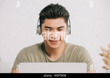 Allegro uomo asiatico utilizzando laptop e ascolto di musica in cuffia Foto Stock