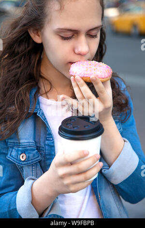 Teen ragazza a mangiare una ciambella di rosa con tazza di caffè la strada al grande città Foto Stock