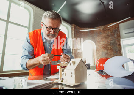 Close up photoshot maschile di architetto e ingegnere fare un modello della futura casa per giovani della famiglia. Uomo che lavora in ufficio con miniatura, disegni, cianografia. Prima Casa, industriale, building concept. Foto Stock