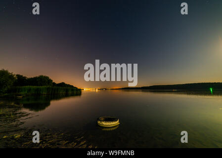 Lago o fiume riva di sabbia con alberi e blu scuro cielo stellato e luce della città sullo sfondo. Carattere tranquillo paesaggio notturno. Foto Stock