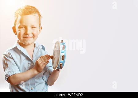 Allegro ragazzo in una maglietta blu tenendo un tamburello e sorridente su uno sfondo luminoso Foto Stock