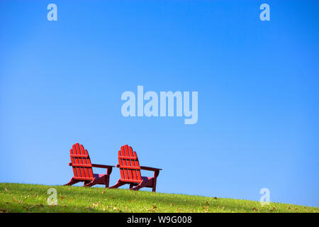 Rosso in legno in stile classico stile Adirondack sedie sono seduti su un verde pendio erboso. Inquadratura orizzontale. Foto Stock