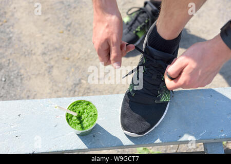 Frullato di verde e acceso - uno stile di vita sano. Vista dettagliata del maschio del runner scarpa sportiva con lacci di legatura su una panchina nel parco per la dieta e perdita di peso concetto per gli uomini. Ottenere pronto per fare jogging e allenamento cardio. Foto Stock