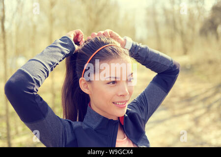 Donna capelli di legatura in coda di cavallo ottenere pronto per l'esecuzione. Bella asiatica giovane adulto di attaccare i suoi lunghi capelli castani su outdoor trail running percorso nel bosco per preparare per l'allenamento. Foto Stock