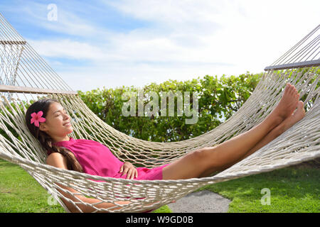 Lunghezza completa di felice rilassata giovane donna sdraiata su un'amaca. Sorridente razza mista Asian / caucasico è femmina in abito. Lei sta godendo del sole nel parco. Foto Stock