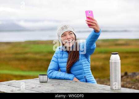 Donna all'aperto utilizzando smart phone tenendo selfie foto su app sullo smartphone mentre seduto fuori indossando warm down jacket. Piuttosto giovane razza mista cinese asiatica donna caucasici in uno stile di vita attivo. Foto Stock