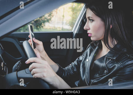 Vista di profilo di giovane donna attraente guardando il suo smartphone durante la guida di un veicolo in un giorno caldo. Driver femminile in giacca di pelle controllo cellulare Foto Stock