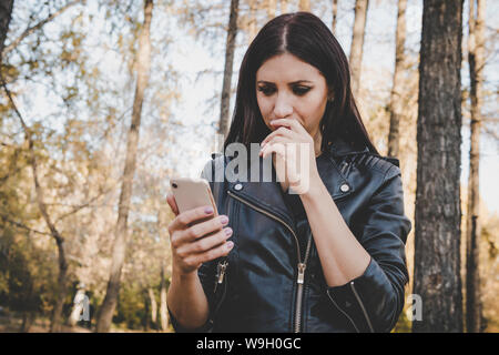 Vista parziale di brunette donna in camicia utilizza lo smartphone in autunno foresta. Passeggiate nel Parco ragazza legge il suo mobile e divenne confuso ottenendo le cattive notizie Foto Stock