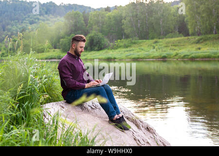 Uomo Barbuto, freelancer seduta con un laptop dal fiume. Il lavoro remoto concetto, freelance Foto Stock