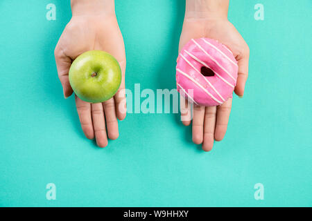 Uno stile di vita sano, cibo e sport concept. Vista dall'alto di una sana versus malsano. Donna mano azienda donut e mela verde blu su sfondo color pastello. Foto Stock