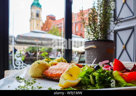 Di salmone al forno con purè di patate, limone, pomodori, cipolle rosse, olive e foglie di insalata su uno sfondo di edifici storici di Torun, Polonia. Resto Foto Stock