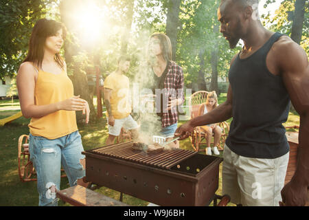 Gruppo di amici felice avente la birra e grigliata alla giornata di sole. Appoggio insieme all'aperto in una radura della foresta o nel cortile. Celebrando e rilassante, laughting. Lo stile di vita di estate, il concetto di amicizia. Foto Stock