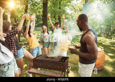 Gruppo di amici felice avente la birra e grigliata alla giornata di sole. Appoggio insieme all'aperto in una radura della foresta o nel cortile. Celebrando e rilassante, laughting. Lo stile di vita di estate, il concetto di amicizia. Foto Stock