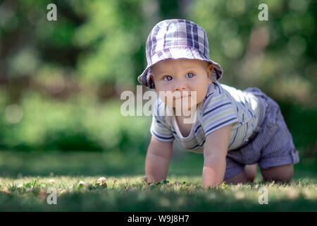 Bambino con bel tappo blu strisciando sul verde del prato in giardino e sorrisi Foto Stock