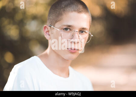 Elegante biondo ragazzo adolescente con gli occhiali e t-shirt bianco in posa sulla natura background closeup. Studente all'esterno. Guardando alla fotocamera. Teenagerhood. Foto Stock