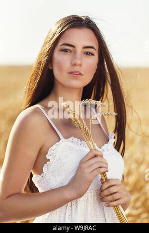 Ritratto di una bellissima ragazza al sole azienda spikelets mature di grano nelle sue mani. Modello in un abito bianco in posa su uno sfondo di un agricolo Foto Stock