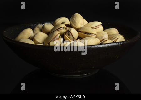 Sacco di tutto mature pistacchio verde scuro in vaso in ceramica isolata sul vetro nero Foto Stock