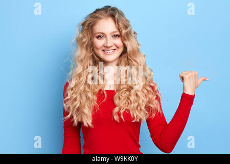 Ottimista giovane donna con toothy sorriso amichevole punti da parte con il pollice, mostra uno spazio vuoto per la vostra pubblicità, isolate su sfondo blu. vendita Foto Stock