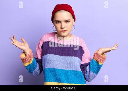 Grave ragazza perplesso solleva entrambe le palme, finge di tenere due elementi, indossa maglia maglione colorato, isolate su sfondo blu. Foto Stock