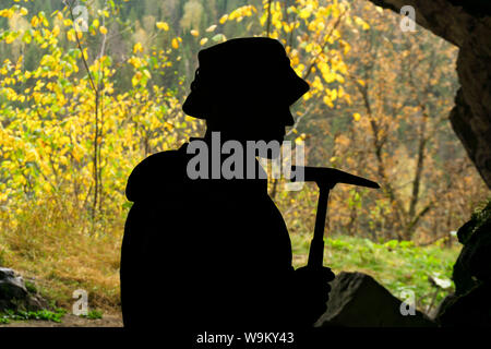 Silhouette di un geologo con un martello geologico in una grotta, sullo sfondo di un ingresso con un luminoso bosco di autunno Foto Stock