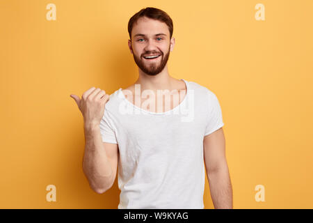 Allegro bello uomo gioioso dispositivo di puntamento con il pollice sulla copyspace dietro la schiena isolata su sfondo giallo. close up verticale. Guarda qui, in questo modo, p Foto Stock