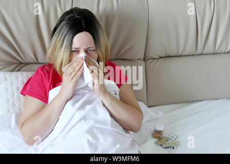 Donna bionda soffia il naso in un fazzoletto di seduta in un letto. Infelice fanciulla soffre di naso che cola, concetto di malati a casa, influenza suina Foto Stock