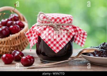 Un vasetto di marmellata di ciliege, cestino di ciliegie, piattino e cucchiaio sul tavolo all'esterno. Foto Stock