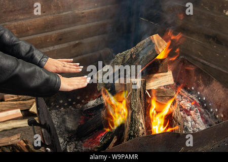 La ragazza si riscalda vicino a fuoco aperto. La donna tiene le mani sopra la fiamma per brindare a se stessa. La masterizzazione di boschi e registri di carbone sono in barbecue grill Foto Stock