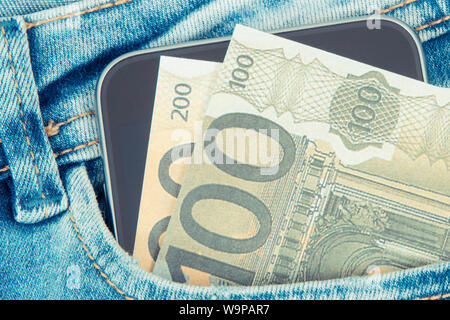 Soldi con il telefono mobile di fronte tasca dei jeans. Concetto di pagare senza contanti o pagamento in contanti per lo shopping Foto Stock