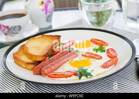 Prima colazione inglese sulla terrazza estiva: uova fritte, salsicce, pomodori e toast. Tazza di caffè. In prossimità della prima colazione in hotel. Foto Stock